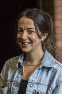 A woman with dark brown hair in a high ponytail wearing a denim jacket with a brick wall behind.