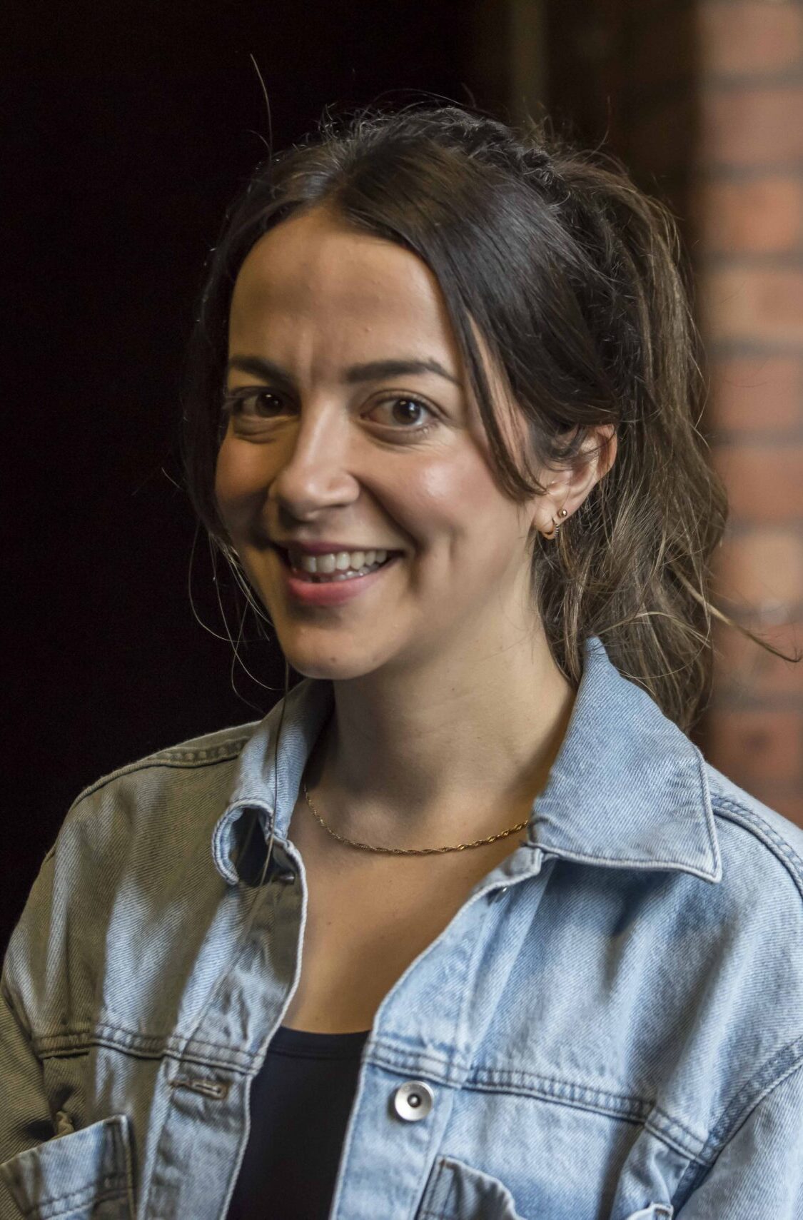 A woman with dark brown hair in a high ponytail wearing a denim jacket with a brick wall behind.
